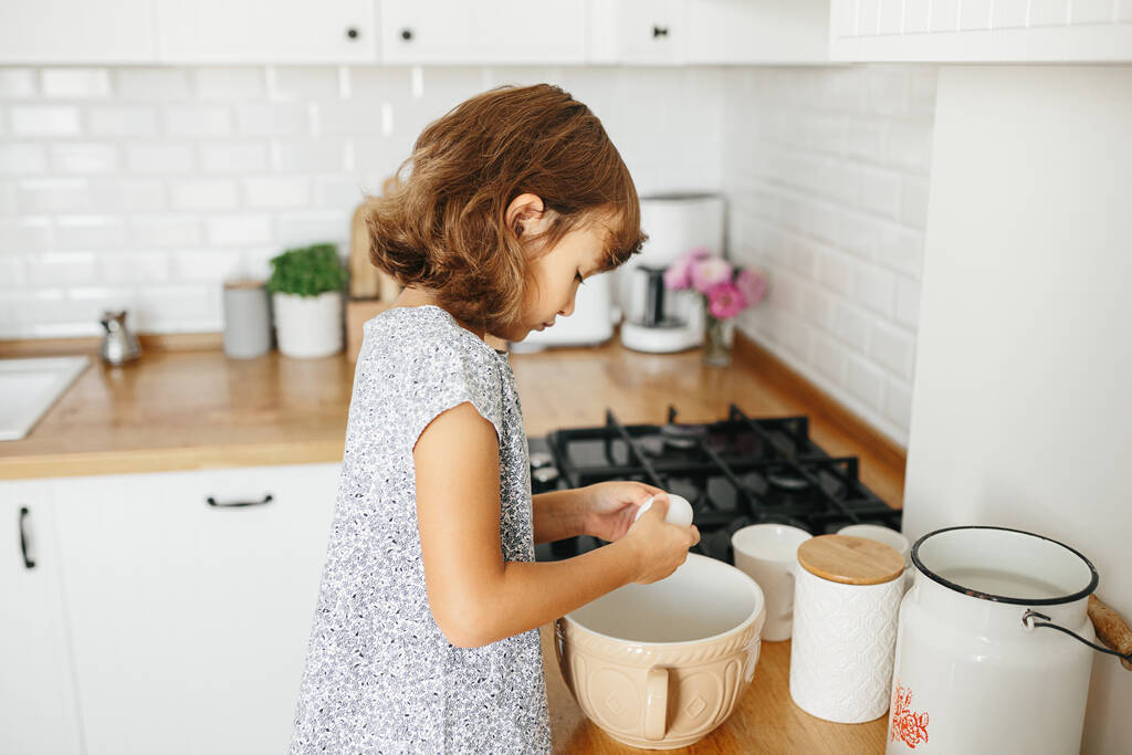 Kindermädchen bereitet Teig zum Backen zu - bricht Ei in Schüssel - Hausmannskost. Konzept der Essenszubereitung, weiße Küche im Hintergrund. Lässige Lifestyle-Fotoserie im echten Leben - Foto, Bild