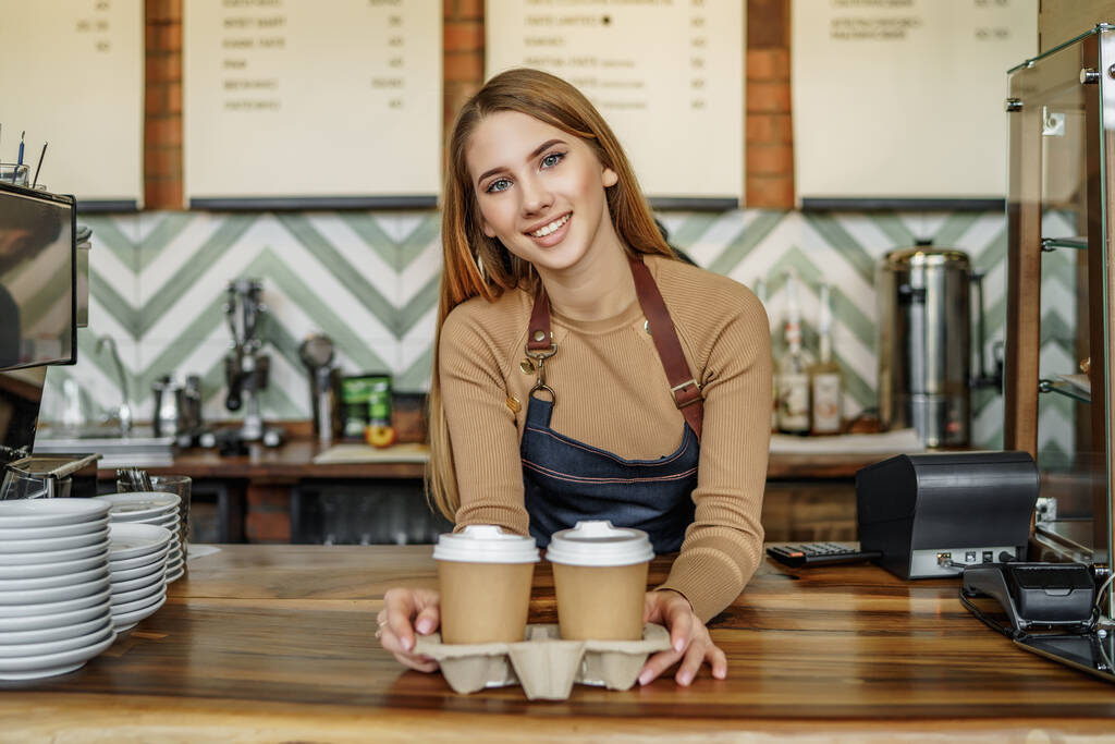 Beautiful blonde female barista is working in coffee shop. Attractive young woman is standing behind the bar counter, making coffee and welcomes customers. - Photo, Image