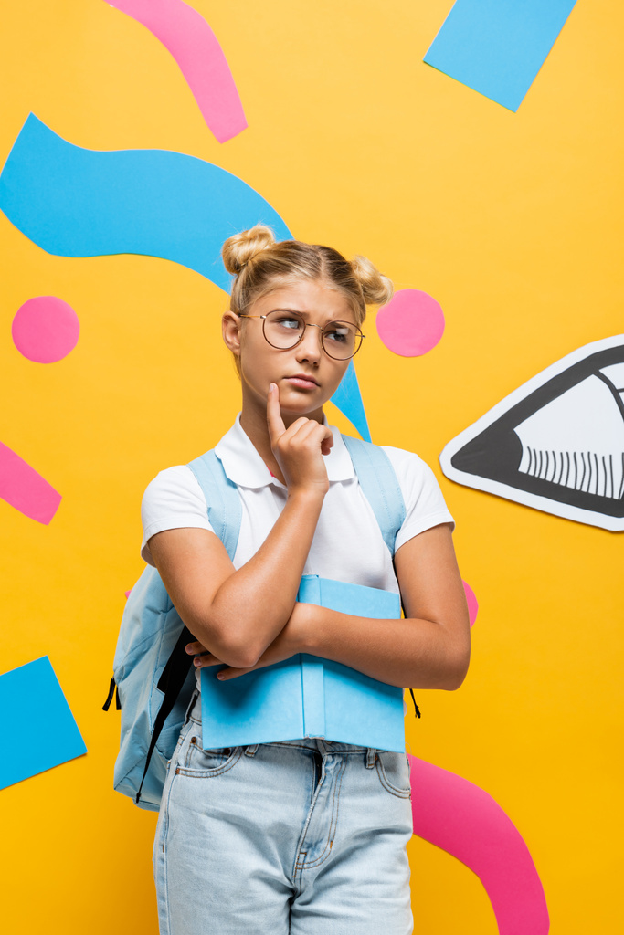 вдумчивая школьница в очках смотрит в сторону, держа книгу возле бумажного карандаша и красочных элементов на желтом - Фото, изображение