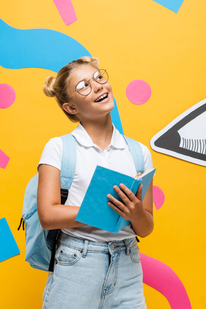 verträumtes Schulkind in Brille, Buch in der Hand und wegschauend auf gelbem Hintergrund mit abstrakten Elementen und Papierstift - Foto, Bild