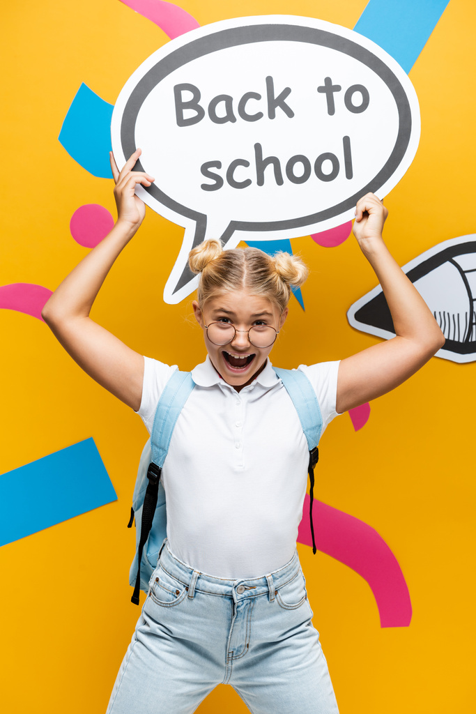 excited schoolkid shouting while holding speech bubble with back to school inscription near paper pencil and abstract elements on yellow - Photo, Image