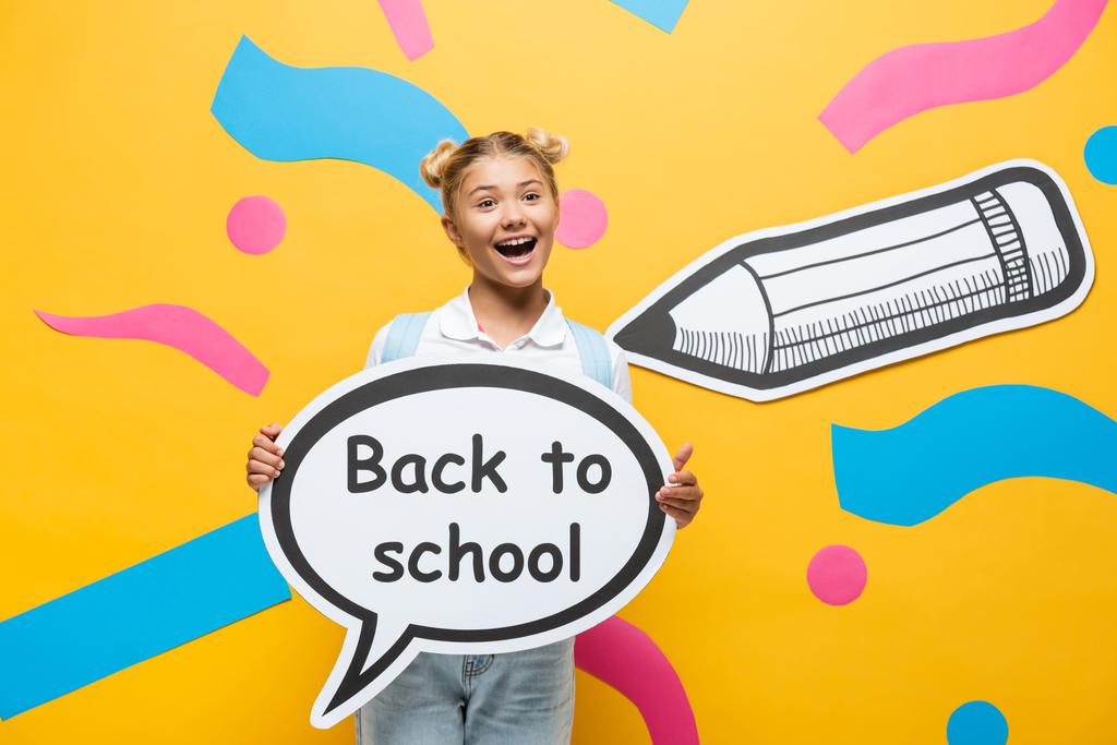 збуджена школярка тримає мовну бульбашку ззаду до школи, що розкладається біля різнокольорових паперових елементів та олівця на жовтому
  - Фото, зображення
