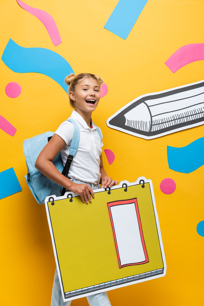 радостный школьник смотрит в камеру, держа макет ксерокса рядом с бумажным карандашом и абстрактными элементами на желтом - Фото, изображение