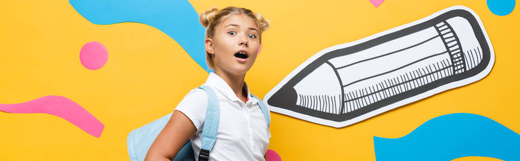 οριζόντια έννοια της κατάπληκτος μαθητής κοιτάζοντας κάμερα σε κίτρινο φόντο με μολύβι χαρτί και πολύχρωμα στοιχεία - Φωτογραφία, εικόνα
