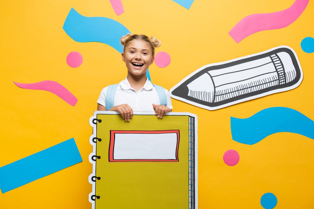 vrolijk schoolmeisje met notebook maquette en kijken naar de camera op gele achtergrond met papier gesneden potlood en decoratieve elementen - Foto, afbeelding