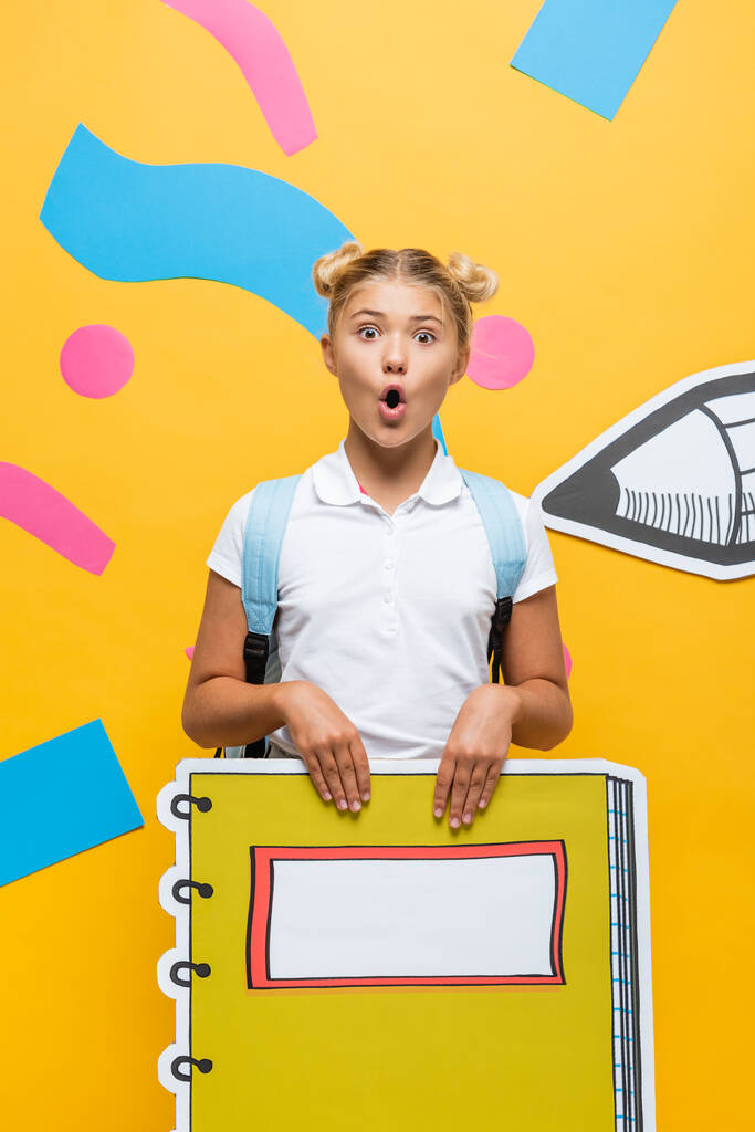 šokované školačka s notebookem maketa při pohledu na kameru na žlutém pozadí s papírem řezané tužkou a barevné prvky - Fotografie, Obrázek