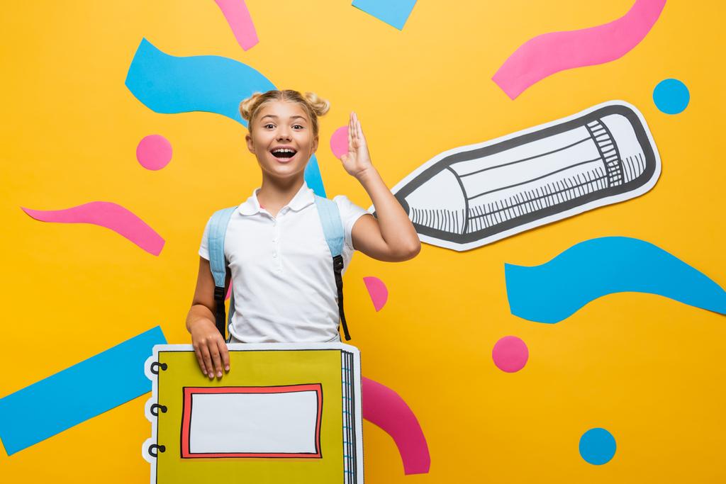 opgewonden schoolkind met notebook maquette heffende hand op gele achtergrond met papier gesneden potlood en veelkleurige elementen  - Foto, afbeelding