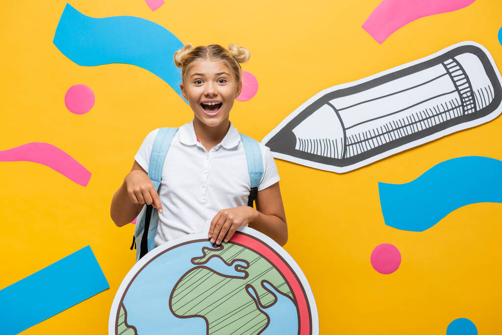 радісний школяр, вказуючи пальцем на макет глобуса біля паперового олівця та різнокольорових елементів на жовтому
 - Фото, зображення