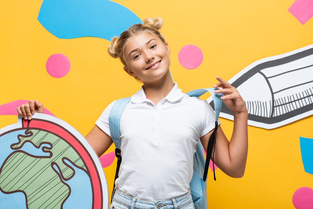 elégedett iskoláslány gazdaság földgömb makett sárga alapon papír ceruza és színes elemek - Fotó, kép