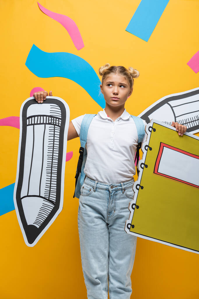 Задумчивый школьник с бумажным карандашом и блокнотом на желтом фоне - Фото, изображение
