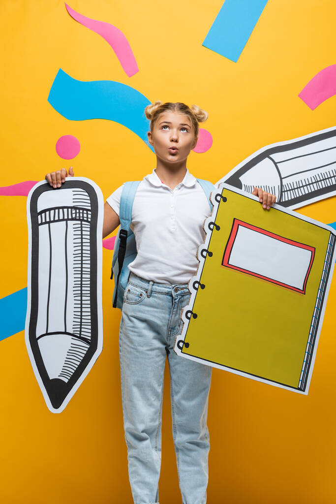 Pensive schoolgirl holding paper pencil and notebook near paper art on yellow background - Photo, Image