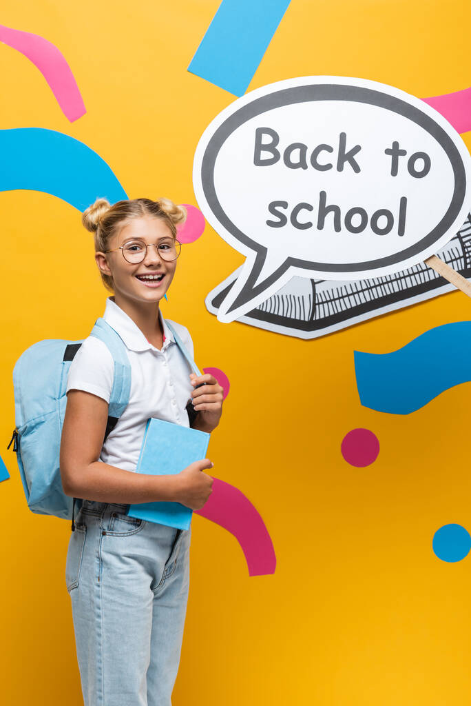 Школа с рюкзаком и книгой, стоящей рядом с речевым пузырём со школьными надписями и бумажными рисунками на жёлтом фоне - Фото, изображение
