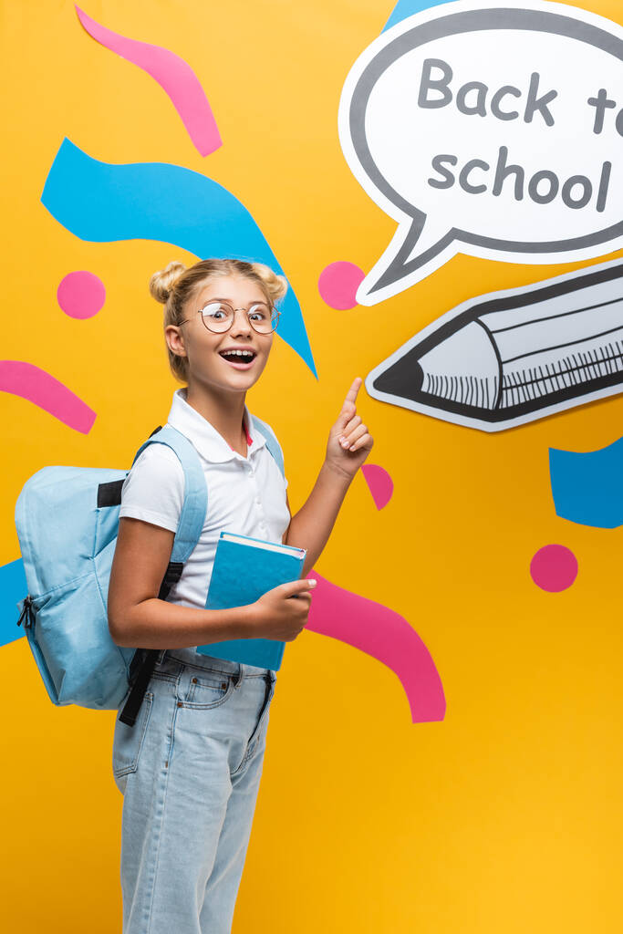 Excited schoolgirl with book pointing at speech bubble wit back to school lettering and paper art on yellow background - Photo, Image