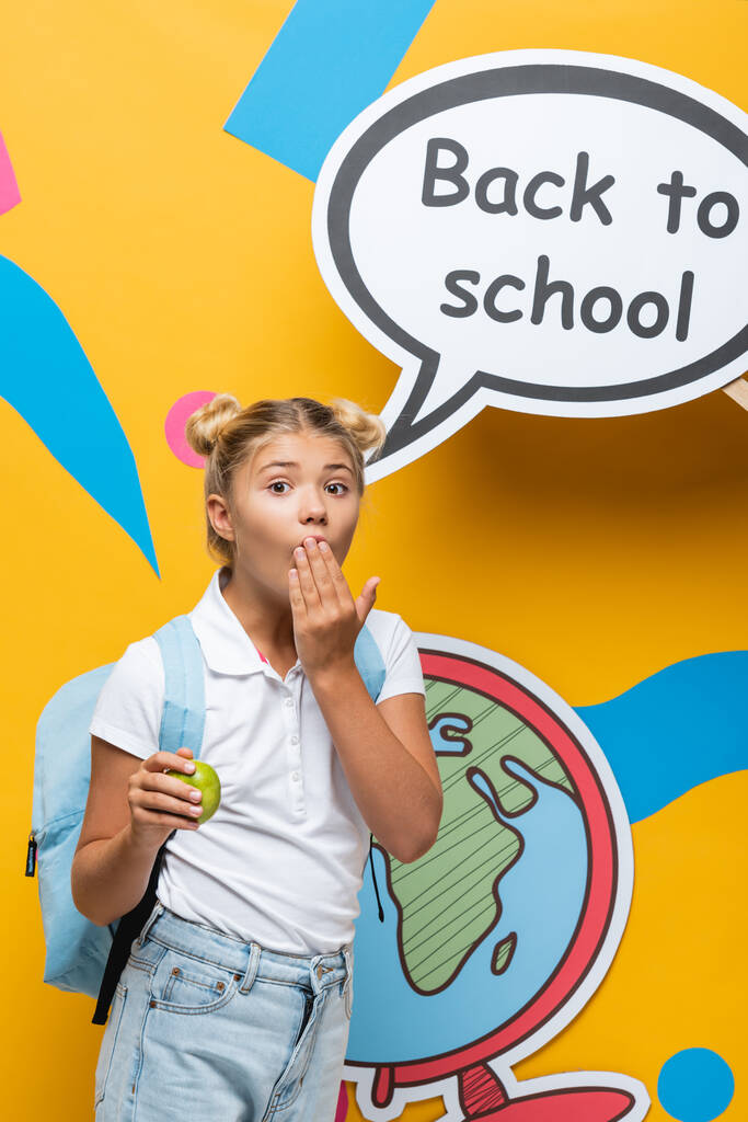 Schockiertes Schulkind hält Apfel und verdeckt Mund in der Nähe der Sprechblase mit Schulbuchstaben auf gelbem Hintergrund - Foto, Bild
