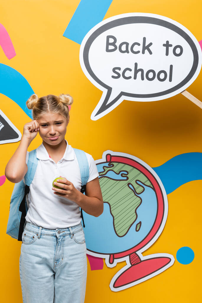 Écolière en pleurs avec pomme debout à côté de l'artisanat du papier et bulle de la parole avec lettrage de retour à l'école sur fond jaune - Photo, image