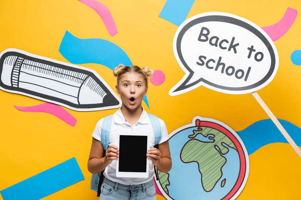 Шокированная школьница держит цифровой планшет рядом с бумажным искусством и мыльным пузырем речи на желтом фоне - Фото, изображение