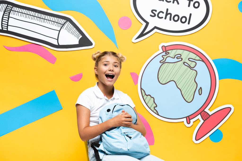 Izgalmas iskoláslány tartó hátizsák mellett beszéd buborék és papír kézműves sárga alapon - Fotó, kép
