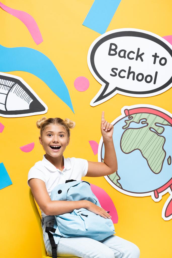 Школьница с рюкзаком, указывающая пальцем на речевой пузырь со школьными надписями и бумажными рисунками на жёлтом фоне - Фото, изображение
