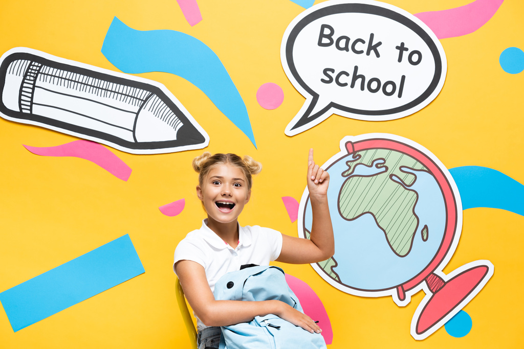 Schoolchild with backpack pointing at back to school lettering and paper elements on yellow background - Photo, Image