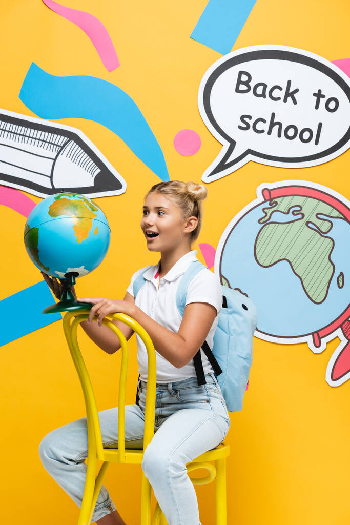 Aufgeregtes Schulkind blickt auf Globus neben Papierbastelei auf gelbem Hintergrund - Foto, Bild