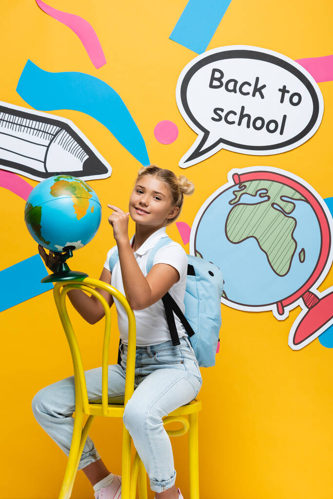 Schulmädchen zeigt auf Globus in der Nähe von Papierkunst mit Schulbuchstaben auf gelbem Hintergrund - Foto, Bild