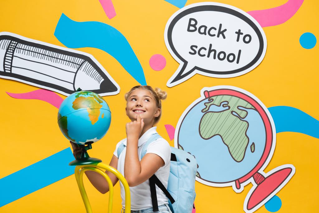 Verträumtes Schulkind mit Globus auf Stuhl neben Papierkunst auf gelbem Hintergrund - Foto, Bild
