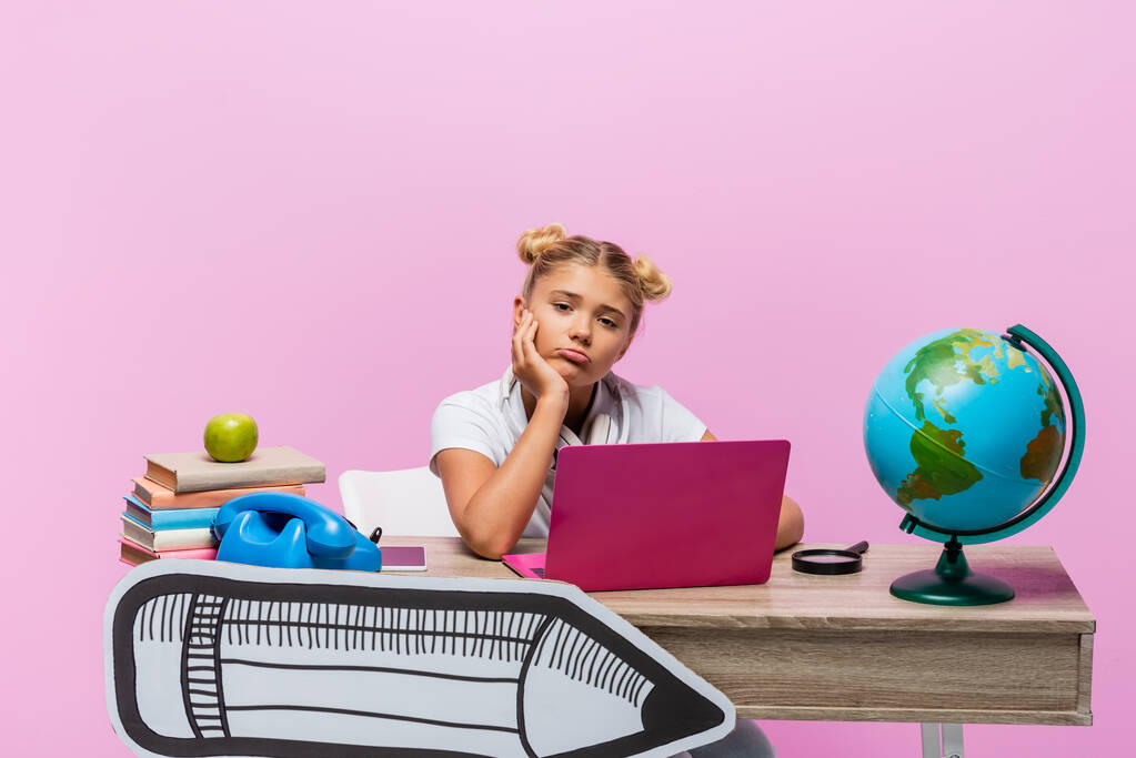 Κουρασμένο κορίτσι κοιτάζοντας κάμερα κοντά στο laptop, τα βιβλία και την τέχνη του χαρτιού στο γραφείο σε ροζ φόντο - Φωτογραφία, εικόνα