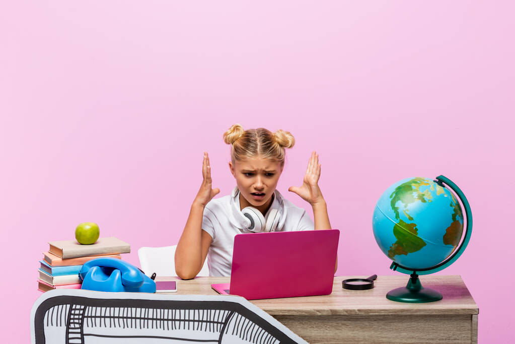 Θυμωμένη κοπέλα με ακουστικά κοιτάζοντας το φορητό υπολογιστή κοντά σε βιβλία και την τέχνη του χαρτιού στο γραφείο απομονώνονται σε ροζ  - Φωτογραφία, εικόνα