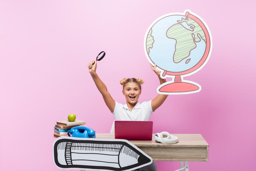 Emozionato scolaro tenendo loupe e globo di carta vicino laptop, cuffie e libri su sfondo rosa - Foto, immagini