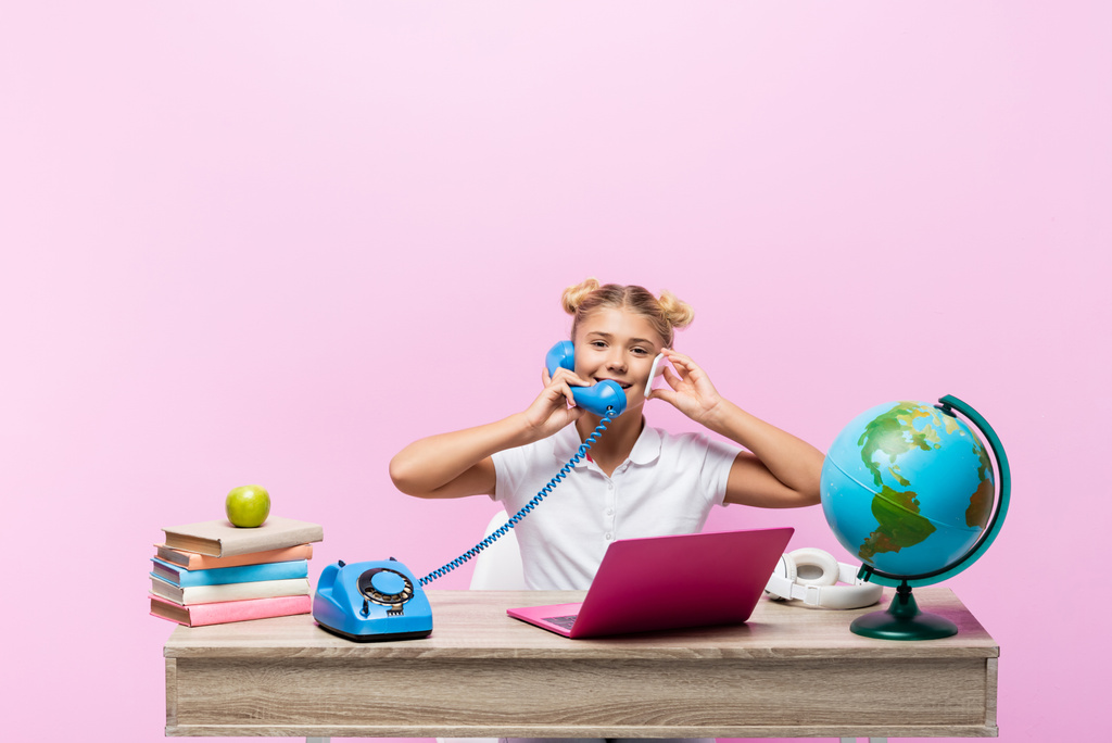 Школярка розмовляє на смартфоні і телефоні біля книг і глобусу ізольовано на рожевому
  - Фото, зображення