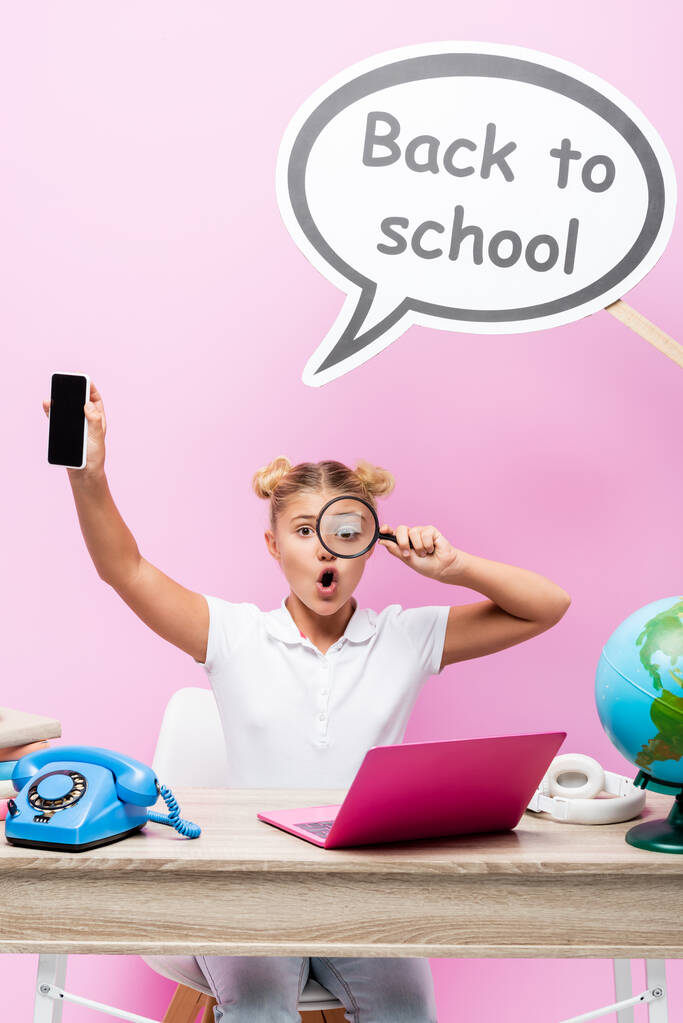Opgewonden schoolmeisje met vergrootglas en smartphone in de buurt van spraakzeepbel, bol en laptop op roze achtergrond - Foto, afbeelding