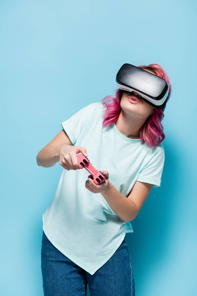 KYIV, UKRAINE - JULY 29, 2020: young woman with pink hair in vr headset playing video game with joystick on blue background - Photo, Image