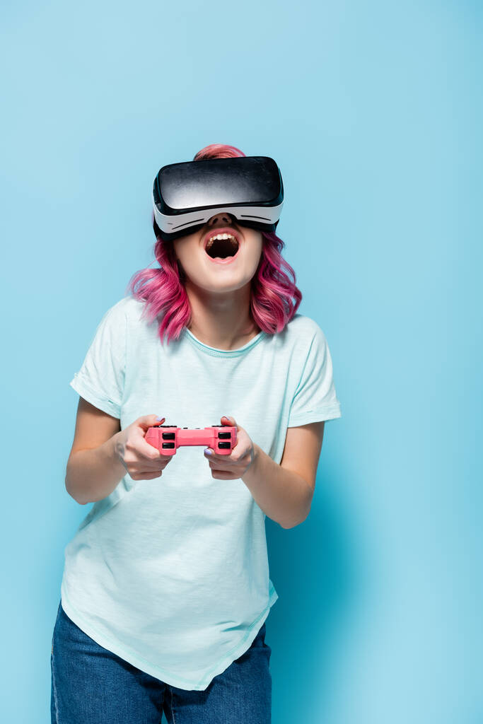 KYIV, UCRANIA - 29 de julio de 2020: mujer joven y excitada con el pelo rosa en los auriculares vr jugando videojuegos con joystick sobre fondo azul - Foto, imagen