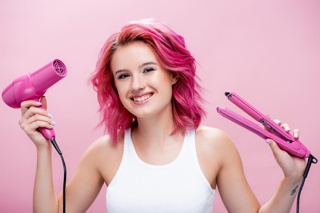 νεαρή γυναίκα με πολύχρωμα μαλλιά κρατώντας ίσιωμα και στεγνωτήρα μαλλιών που απομονώνονται σε ροζ - Φωτογραφία, εικόνα