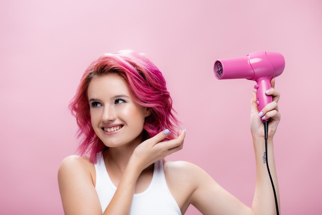 młoda kobieta z kolorowymi włosami trzyma suszarkę do włosów i uśmiecha się odizolowany na różowy - Zdjęcie, obraz