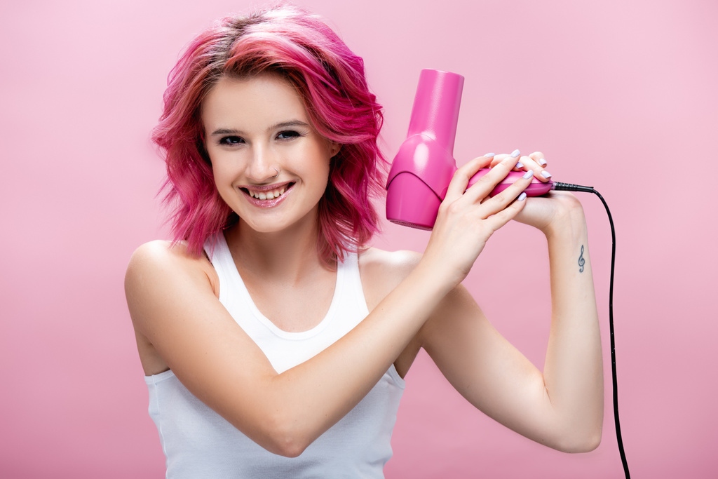 giovane donna con capelli colorati in possesso di asciugacapelli e sorridente isolato su rosa - Foto, immagini
