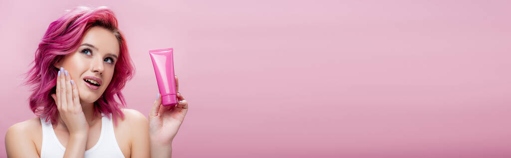 jeune femme rêveuse aux cheveux colorés tenant tube de crème cosmétique isolé sur rose, plan panoramique - Photo, image