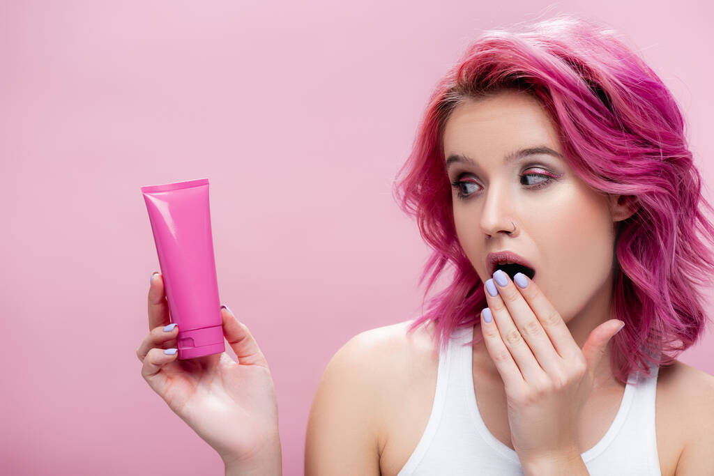 zszokowana młoda kobieta z kolorowymi włosami patrząca na tubę kremu kosmetycznego odizolowanego na różowo - Zdjęcie, obraz