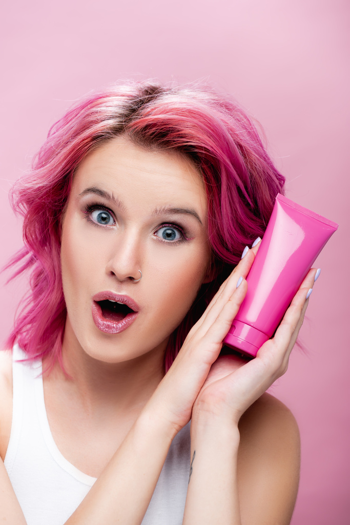 surpris jeune femme avec des cheveux colorés tenant tube de crème cosmétique isolé sur rose - Photo, image