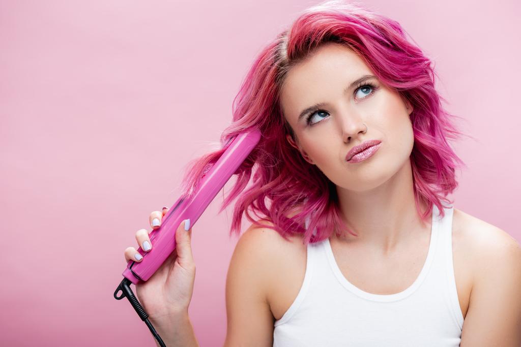verträumte junge Frau mit buntem Haar mit Glätteisen isoliert auf rosa - Foto, Bild