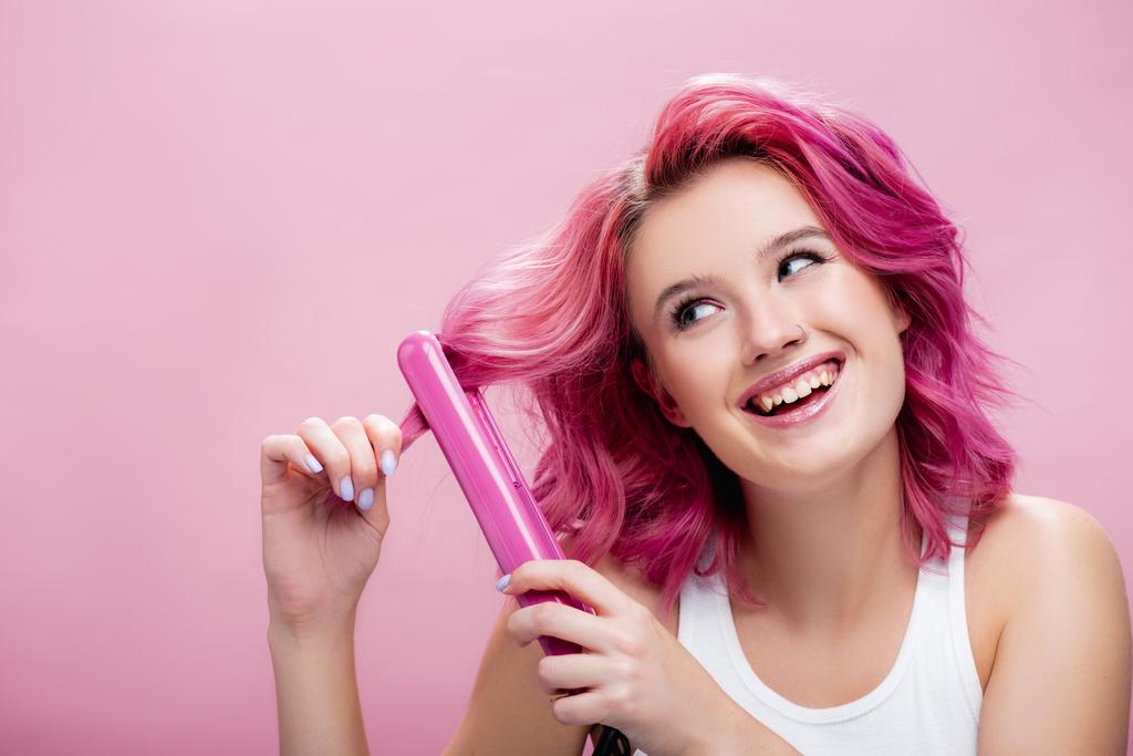 νεαρή γυναίκα με πολύχρωμα μαλλιά χρησιμοποιώντας ίσιωμα απομονώνονται σε ροζ - Φωτογραφία, εικόνα