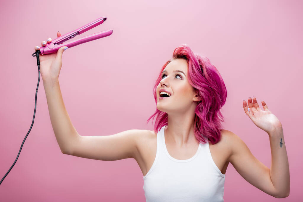 νεαρή γυναίκα με πολύχρωμα μαλλιά κοιτάζοντας straightener απομονώνονται σε ροζ - Φωτογραφία, εικόνα
