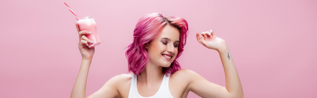 young woman with colorful hair holding strawberry milkshake in glass with drinking straw and dancing isolated on pink, panoramic shot - Photo, Image