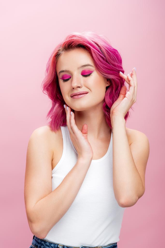 jovem com cabelos coloridos e maquiagem posando com as mãos perto do rosto e olhos fechados isolados no rosa - Foto, Imagem
