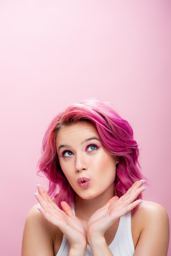 mujer joven sorprendida con el pelo colorido y maquillaje posando con las manos cerca de la cara aislada en rosa - Foto, imagen