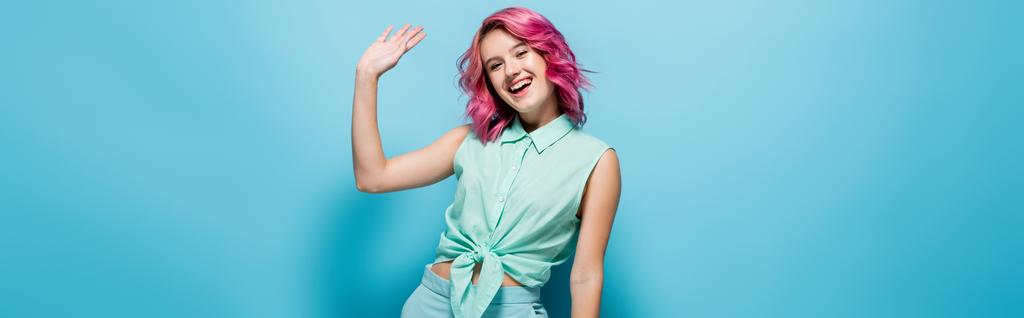mladá žena s růžové vlasy mává rukou na modrém pozadí, panoramatický výstřel - Fotografie, Obrázek