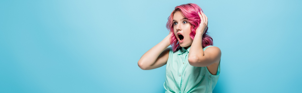 σοκαρισμένη νεαρή γυναίκα με ροζ μαλλιά και ανοιχτό στόμα κρατώντας το κεφάλι σε μπλε φόντο, πανοραμική βολή - Φωτογραφία, εικόνα