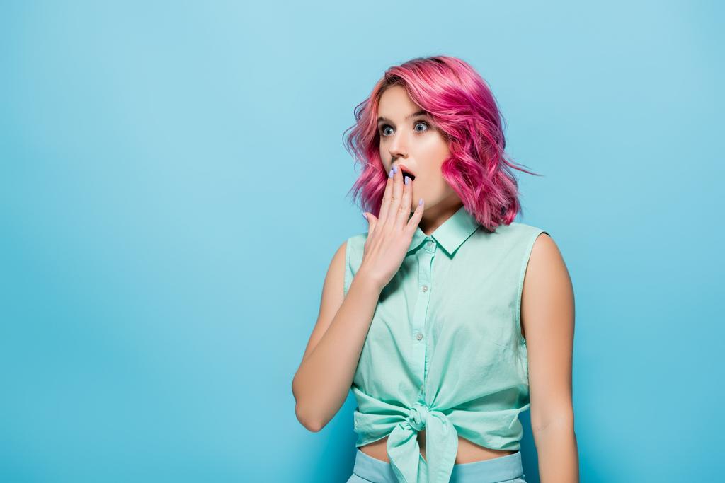 σοκαρισμένη νεαρή γυναίκα με ροζ μαλλιά που καλύπτει το στόμα σε μπλε φόντο - Φωτογραφία, εικόνα