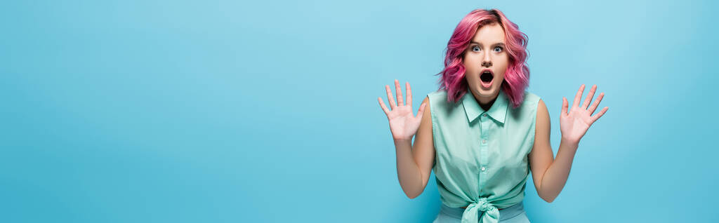 shocked young woman with pink hair and open mouth showing hands on blue background, panoramic shot - Photo, Image