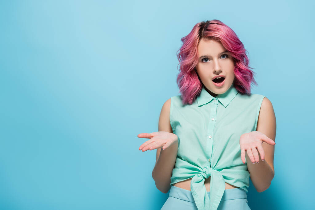 surprised young woman with pink hair and open mouth showing shrug gesture on blue background - Photo, Image
